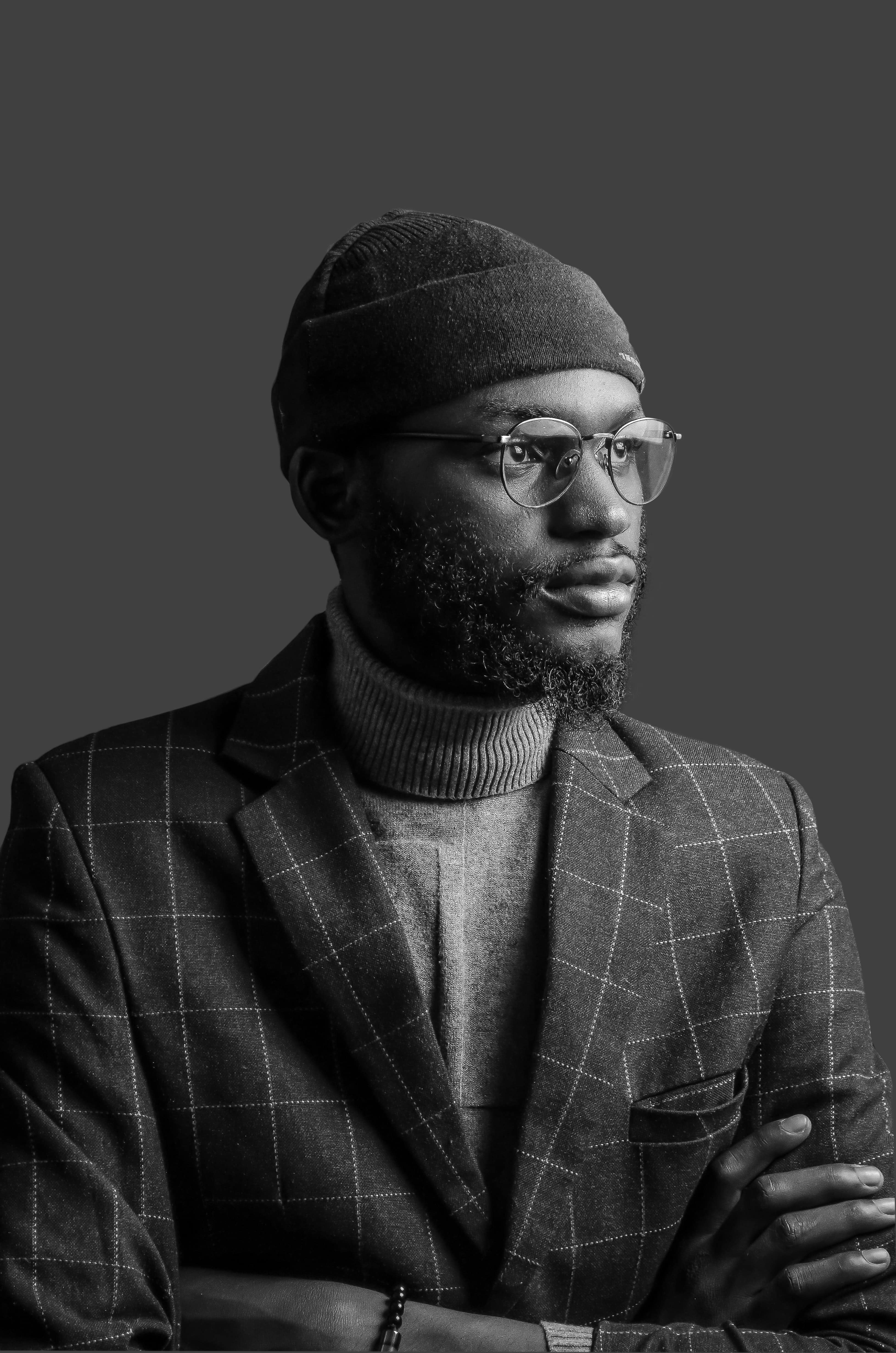 Black and white portrait of male wearing glasses, wool cap, sweater and a jacket.